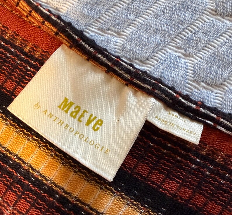 Puede incluir: Primer plano de un textil con una etiqueta que dice "Maeve by Anthropologie". El textil presenta un estampado de rayas en tonos naranja, amarillo y negro. La etiqueta tambi&eacute;n indica la talla XS y que est&aacute; hecho en Turqu&iacute;a.