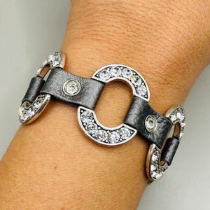 May include: A silver and gray bracelet with clear crystal accents. The bracelet features a series of silver-toned, open-circle links, each encrusted with clear crystals. The links are connected by gray leather straps.