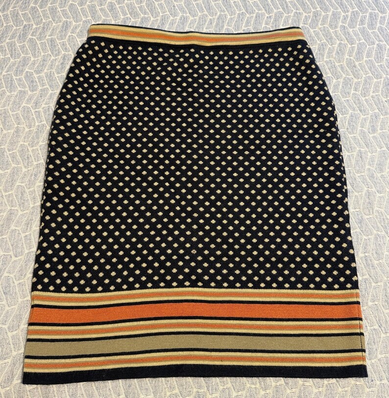 May include: A navy blue pencil skirt with a white polka dot pattern. The skirt has a striped design in orange, tan, and navy blue at the hem and waistband. The skirt is made of a knit fabric.