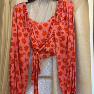 K&ouml;nnte beinhalten: Eine rosa Bluse mit langen &Auml;rmeln und rotem Blumendruck. Die Bluse hat einen Knoten in der Taille und einen quadratischen Ausschnitt.