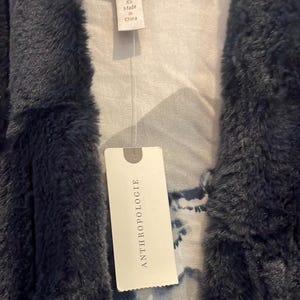 Op de afbeelding: Een donkerblauwe nep bontjas met een wit label waarop "ANTHROPOLOGIE" staat en een kleiner wit label waarop "XS Made in China" staat.