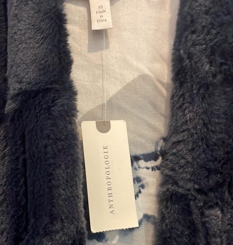 Op de afbeelding: Een donkerblauwe nep bontjas met een wit label waarop "ANTHROPOLOGIE" staat en een kleiner wit label waarop "XS Made in China" staat.