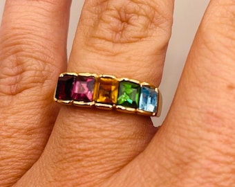May include: A gold band ring with five square-shaped gemstones. The gemstones are red, orange, green, blue, and light blue.