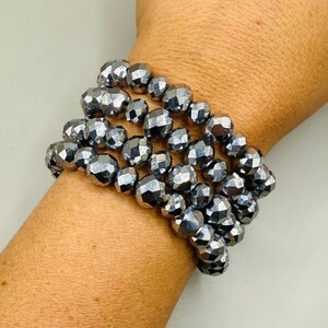 May include: A stack of three bracelets made of faceted, metallic gray beads. The beads are round and have a reflective, shiny surface. The bracelets are worn on a wrist, showcasing the jewelry's design and color.
