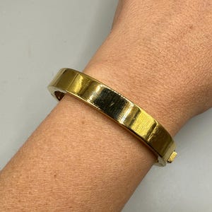 以下が含まれることがあります： ゴールドカラーのヒンジ付きバングルブレスレット。ブレスレットは長方形で、滑らかで反射する表面を持っています。ブレスレットは手首に着用されています。
