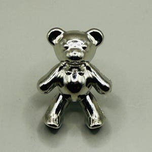 May include: A small, silver-colored teddy bear figurine. The bear has a shiny, reflective surface and is standing upright with its arms and legs extended. The bear's features include small, rounded ears and a detailed face.