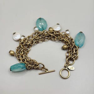 May include: Gold-tone charm bracelet featuring a toggle clasp. The bracelet has multiple strands of chain and is adorned with blue, white, and gold-colored beads. The bracelet has a JE tag.