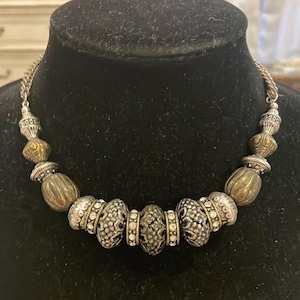 May include: A silver-toned necklace with ornate beads and accents. The necklace features a variety of bead shapes, including round, oval, and textured designs. Some beads are embellished with clear crystals. The necklace is displayed on a black mannequin.