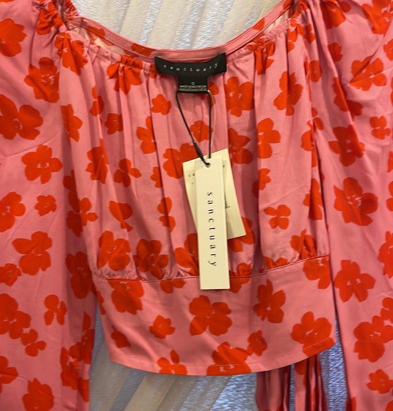 K&ouml;nnte beinhalten: Eine rosa Bluse mit langen &Auml;rmeln und rotem Blumendruck. Die Bluse hat einen quadratischen Ausschnitt und eine Schleife in der Taille. Das Etikett auf der Bluse lautet "sanctuary".