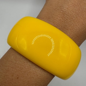 May include: A wide, solid yellow bangle bracelet with a smooth, glossy finish. The bracelet features a curved, rectangular shape with rounded edges and a reflective surface that catches light, creating a bright, uniform appearance on the wrist.