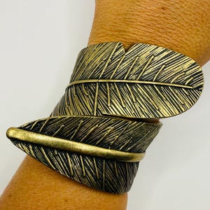 May include: A wrap-around cuff bracelet crafted from antique brass-toned metal, featuring a detailed design of two overlapping leaves with prominent veins and textured surfaces that curve around the wrist in a continuous, layered form.
