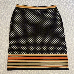 May include: A black pencil skirt with a white polka dot pattern. The skirt has a striped border in shades of orange, tan, and black at the hem. The waistband has a thin stripe of orange and tan.