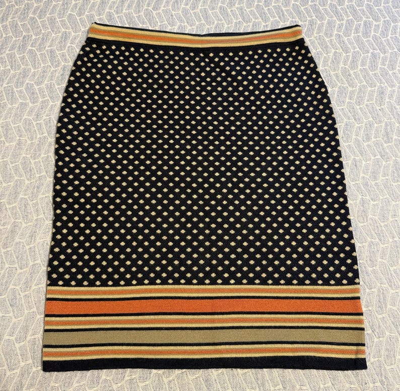 May include: A black pencil skirt with a white polka dot pattern. The skirt has a striped border in shades of orange, tan, and black at the hem. The waistband has a thin stripe of orange and tan.
