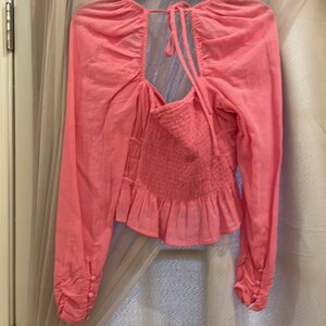 May include: A pink long-sleeved blouse with a gathered neckline, a tie at the back, and a ruffled hem.