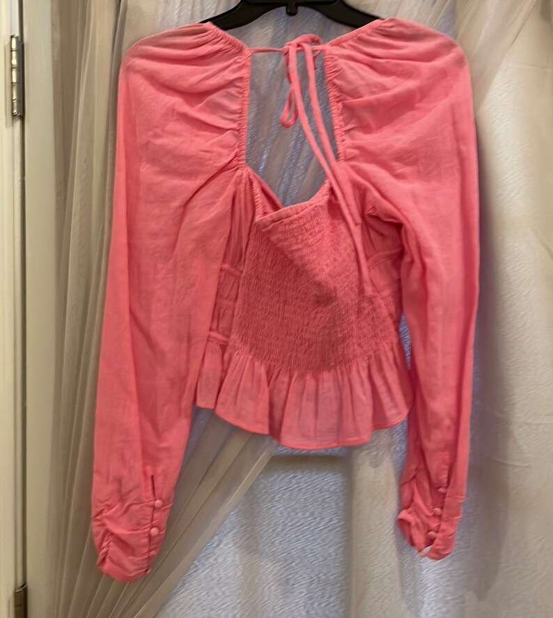 May include: A pink long-sleeved blouse with a gathered neckline, a tie at the back, and a ruffled hem.