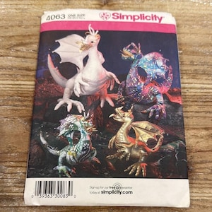 May include: Simplicity pattern 4063 featuring four dragon designs. The dragons are in various colors, including white, gold, and iridescent hues. The pattern includes the text "One Size" and "Sign-up for our free newsletter today at simplicity.com."