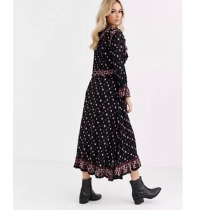 May include: A black midi dress with a white polka dot pattern and a pink floral trim. The dress has a ruffled neckline and long sleeves. The model is wearing black ankle boots.