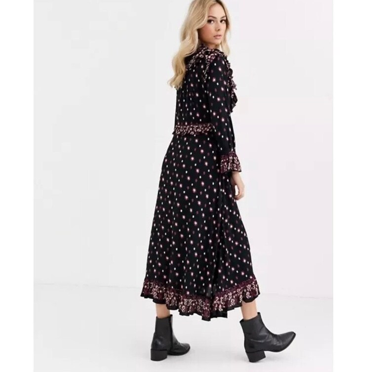 May include: A black midi dress with a white polka dot pattern and a pink floral trim. The dress has a ruffled neckline and long sleeves. The model is wearing black ankle boots.