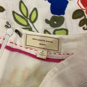 May include: White linen dress with embroidered floral details in green, yellow, blue, and red. The dress has a pink waistband and a label that reads "Moulinette Soeurs vetements" and "Made in India Fabrique en Inde".