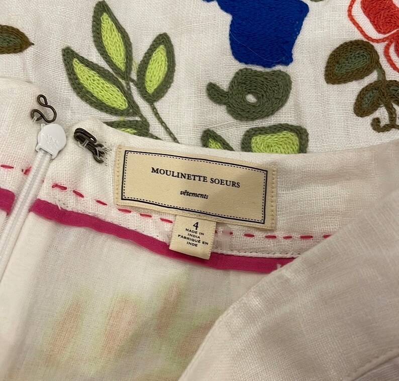May include: White linen dress with embroidered floral details in green, yellow, blue, and red. The dress has a pink waistband and a label that reads "Moulinette Soeurs vetements" and "Made in India Fabrique en Inde".
