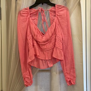 May include: A pink, long-sleeved blouse with a ruffled hem and a tie at the back of the neck.