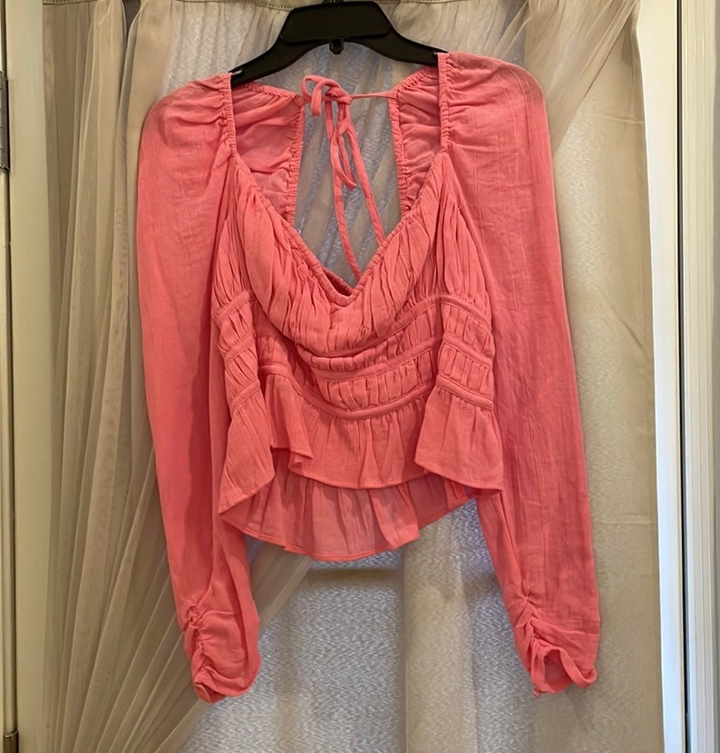 May include: A pink, long-sleeved blouse with a ruffled hem and a tie at the back of the neck.