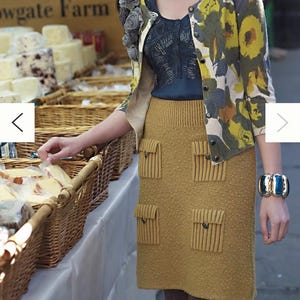 May include: A woman wearing a mustard yellow knitted skirt with multiple pockets and a floral cardigan. The skirt has a textured surface and the cardigan has a white background with yellow, green, and blue floral accents.