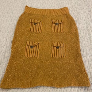 May include: A mustard yellow knitted skirt with four large patch pockets, each with a button closure.