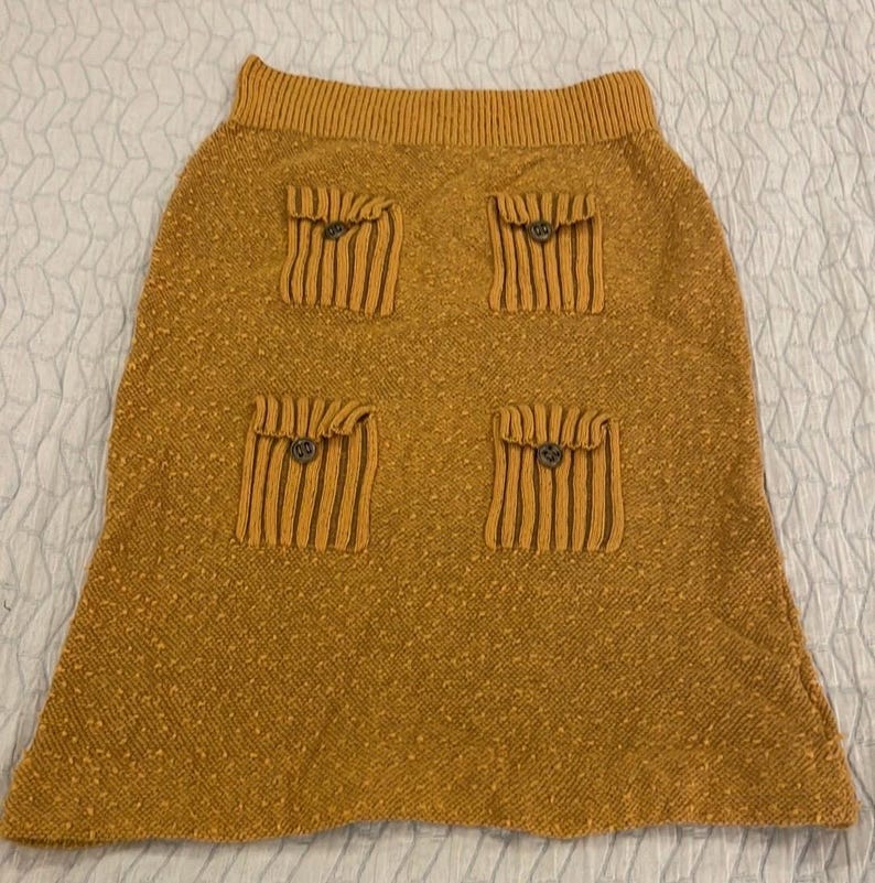 May include: A mustard yellow knitted skirt with four large patch pockets, each with a button closure.