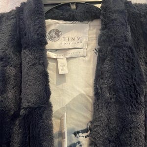 Op de afbeelding: Zwarte nep bontjas met een witte en grijze geprinte voering. De jas heeft een label met de tekst "Tiny Editions" en "Anthropologie". Het label geeft ook aan dat de jas maat XS heeft en gemaakt is in China.