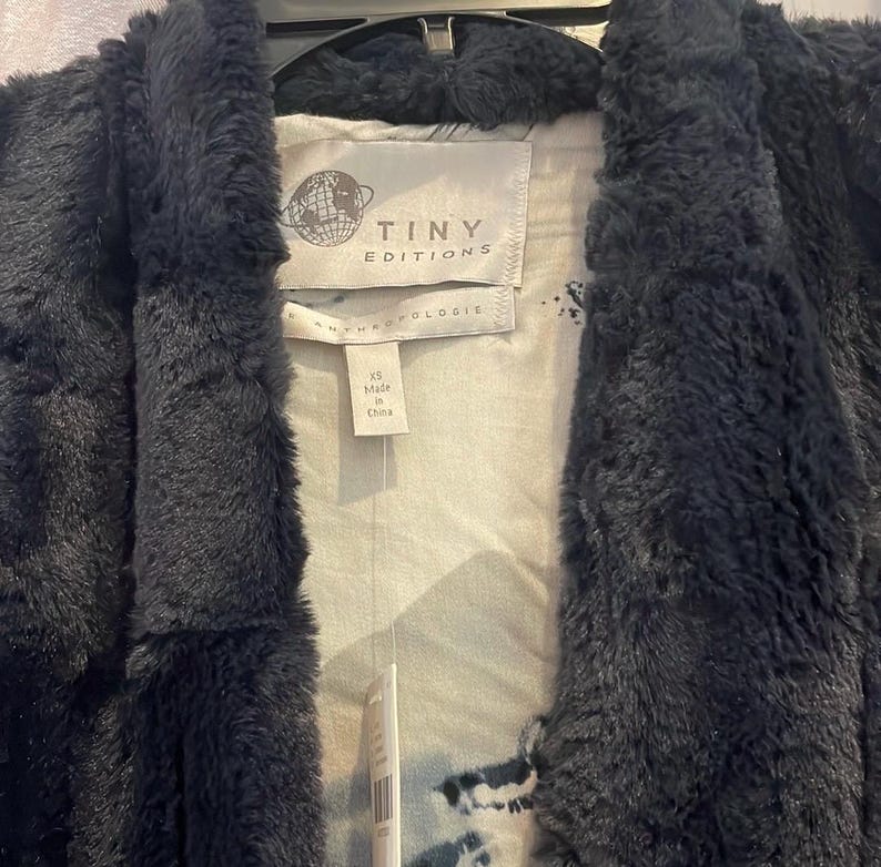 Op de afbeelding: Zwarte nep bontjas met een witte en grijze geprinte voering. De jas heeft een label met de tekst "Tiny Editions" en "Anthropologie". Het label geeft ook aan dat de jas maat XS heeft en gemaakt is in China.