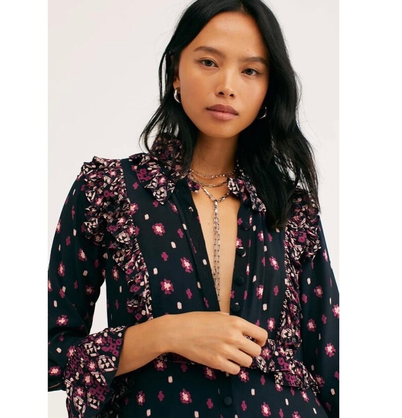 May include: A black and pink patterned long-sleeved shirt with a ruffled collar and cuffs. The shirt has a button-up front and a relaxed fit.