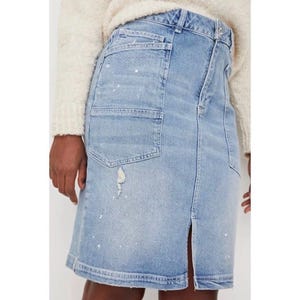 May include: Light blue denim skirt with a front slit and a distressed look. The skirt features a large pocket on the front and white paint splatters. The model is wearing a cream-colored sweater.
