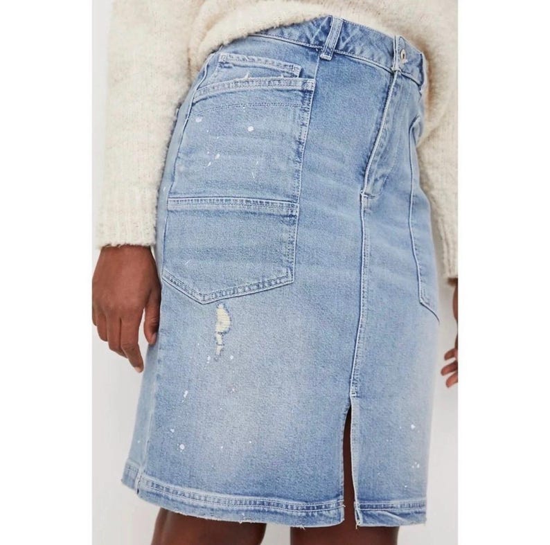 May include: Light blue denim skirt with a front slit and a distressed look. The skirt features a large pocket on the front and white paint splatters. The model is wearing a cream-colored sweater.