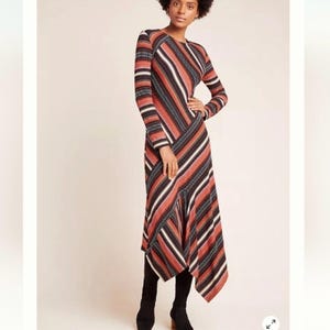 Puede incluir: Un vestido de manga larga con un estampado de rayas en tonos &oacute;xido, negro y blanco. El vestido tiene un cuello redondo y un dobladillo asim&eacute;trico. La modelo lleva botas negras.