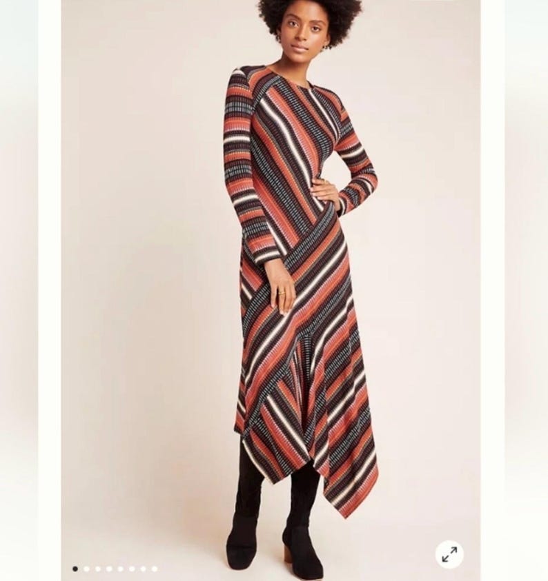 Puede incluir: Un vestido de manga larga con un estampado de rayas en tonos &oacute;xido, negro y blanco. El vestido tiene un cuello redondo y un dobladillo asim&eacute;trico. La modelo lleva botas negras.