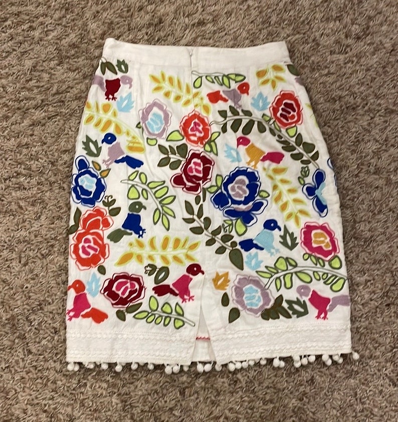 May include: A white skirt with a colorful floral and bird embroidery pattern. The skirt has a white pom-pom trim around the bottom.