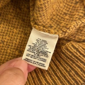 May include: A close-up of a mustard yellow sweater with a white tag that reads "77% Wool, 20% Cotton, 3% Nylon, 1% Spandex. Hand wash cold. Do not bleach. Do not wring. Only non-chlorine bleach as needed. Dry flat. Low iron as needed. May be dry cleaned. French on reverse."
