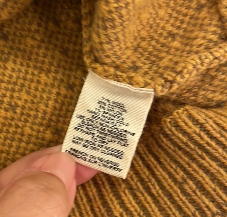 May include: A close-up of a mustard yellow sweater with a white tag that reads "77% Wool, 20% Cotton, 3% Nylon, 1% Spandex. Hand wash cold. Do not bleach. Do not wring. Only non-chlorine bleach as needed. Dry flat. Low iron as needed. May be dry cleaned. French on reverse."