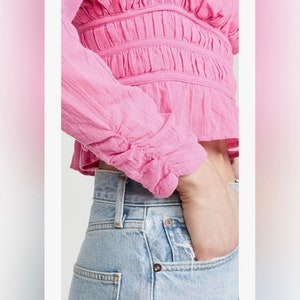 May include: A pink, long-sleeved, cropped blouse with a ruffled neckline and cinched waist. The blouse has a gathered, textured fabric and is worn with light blue jeans.