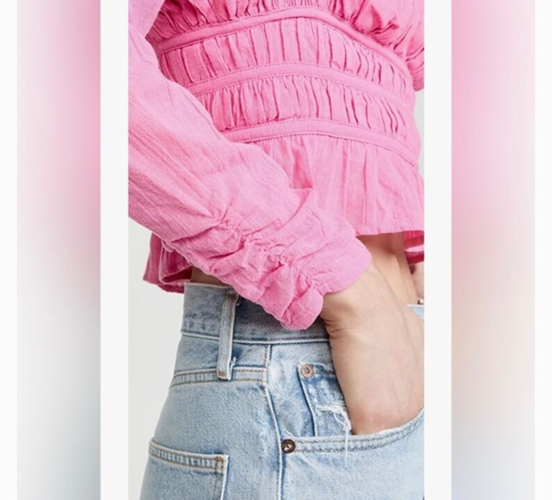 May include: A pink, long-sleeved, cropped blouse with a ruffled neckline and cinched waist. The blouse has a gathered, textured fabric and is worn with light blue jeans.