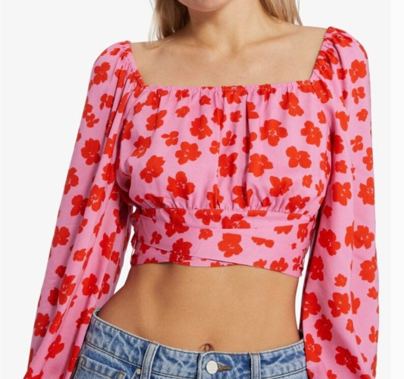 K&ouml;nnte beinhalten: Ein rosa-rotes Oberteil mit Blumenmuster, langen &Auml;rmeln und quadratischem Ausschnitt. Das Oberteil hat einen G&uuml;rtel in der Taille.
