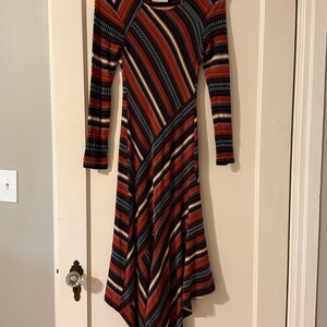 Puede incluir: Un vestido de manga larga con un patr&oacute;n de rayas diagonales &uacute;nico en rojo, negro, blanco y beige. El vestido tiene un escote redondo y un dobladillo asim&eacute;trico acampanado. La tela parece ser de punto acanalado.