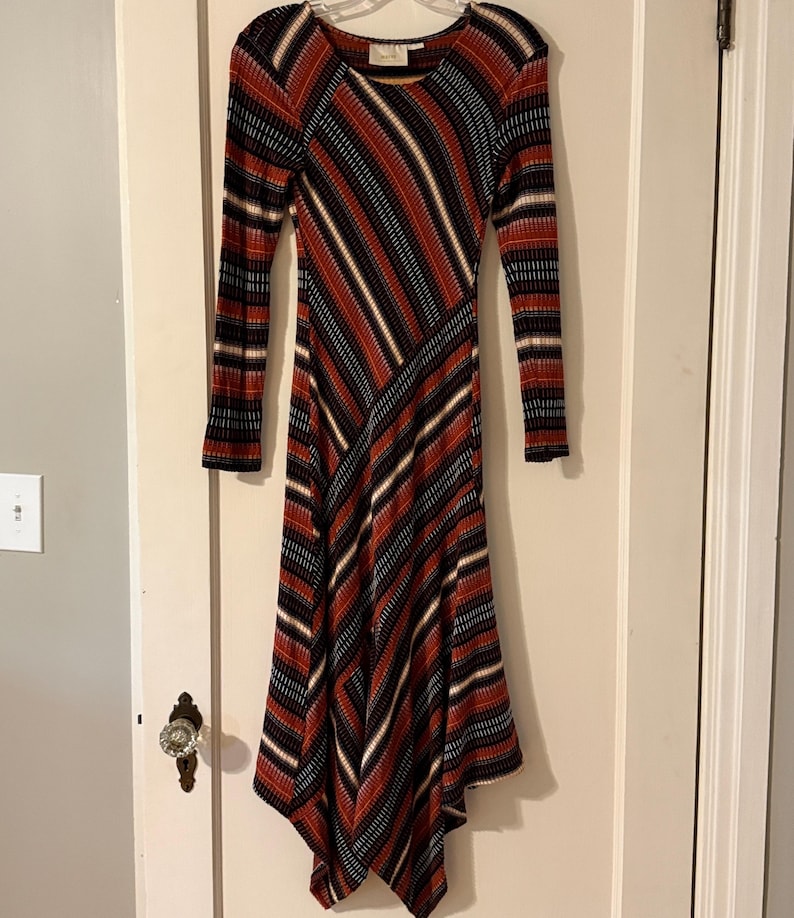 Puede incluir: Un vestido de manga larga con un patr&oacute;n de rayas diagonales &uacute;nico en rojo, negro, blanco y beige. El vestido tiene un escote redondo y un dobladillo asim&eacute;trico acampanado. La tela parece ser de punto acanalado.
