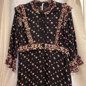 May include: A black long-sleeved dress with a white and pink floral print. The dress has a ruffled collar and cuffs, and a button-up front.