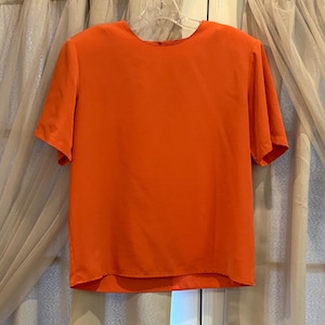 May include: Bright orange short-sleeved blouse. The top has a round neckline and a loose fit. The fabric appears to be a lightweight material, suitable for warmer weather. The blouse is hanging on a clear hanger.