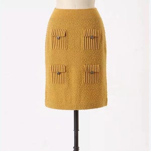 May include: A mustard yellow knitted skirt with four decorative pockets and buttons. The skirt is on a mannequin.