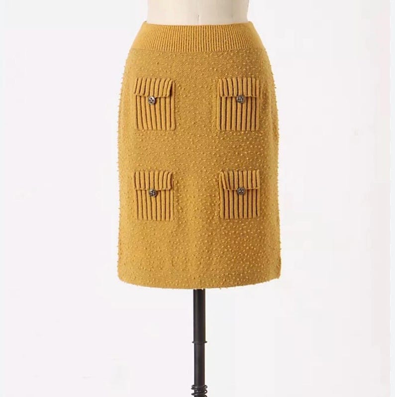 May include: A mustard yellow knitted skirt with four decorative pockets and buttons. The skirt is on a mannequin.
