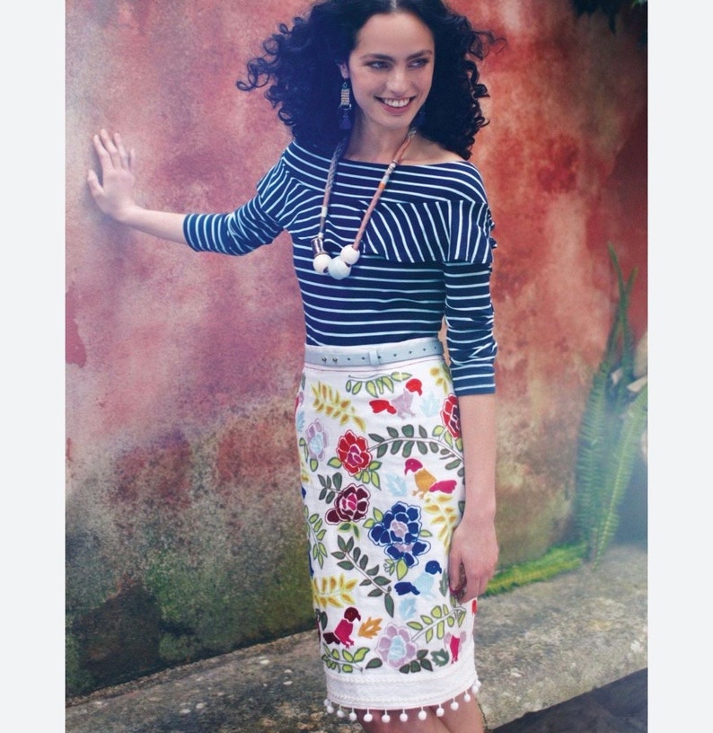 May include: A woman wearing a blue and white striped off-the-shoulder top with a white beaded necklace and a white skirt with colorful embroidered flowers and birds. The skirt has a light blue belt and a white pom-pom trim.