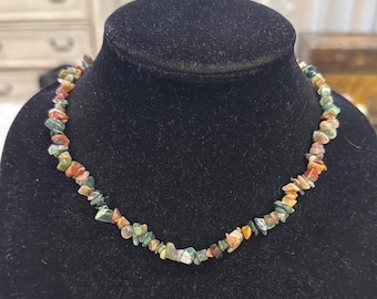 May include: A necklace made of small, irregularly shaped stones in shades of green, orange, and beige. The necklace is displayed on a black velvet bust, showcasing the natural colors and textures of the gemstones.